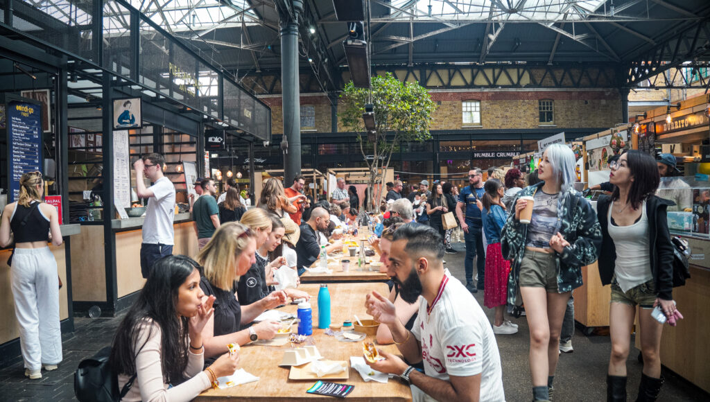 Communal dining tables – Spitalfields food court – Diverse diners eating street‑food lunches at long wooden tables beneath iron‑and‑glass roof.