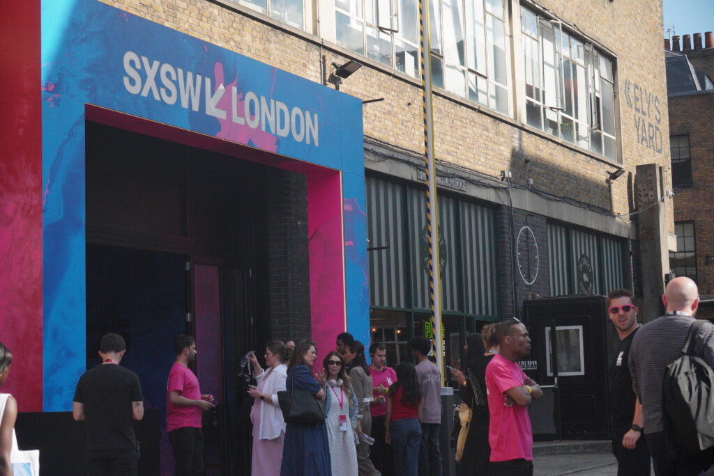 SXSW London entrance – Brick Lane – Visitors queuing beneath cyan‑and‑magenta "SXSW London" archway on Brick Lane.
