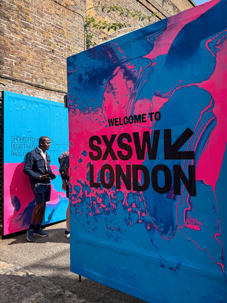 SXSW London welcome cube – Blue‑and‑pink art installation cube reading “Welcome to SXSW London” in Truman Brewery yard, East London street art backdrop.