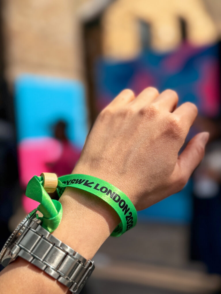 SXSW London wristband close‑up – Hand wearing green “SXSW London 2025” fabric wristband, colourful festival mural blurred in background.