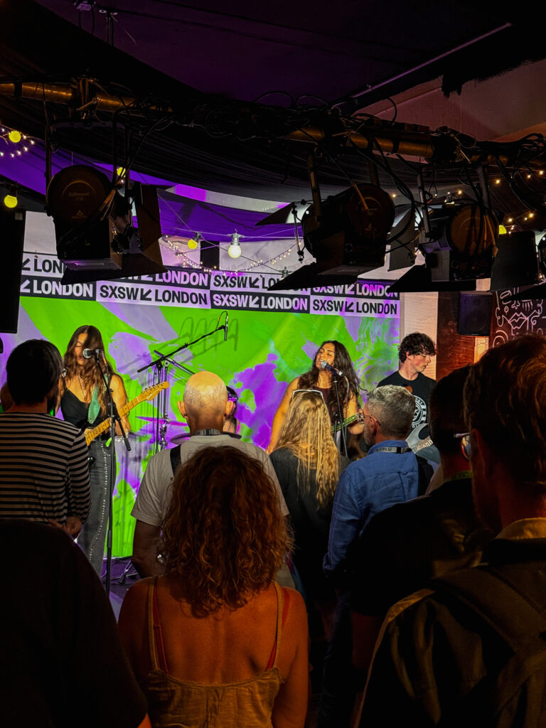 Indie gig at SXSW London – club stage – Small venue crowd watching psychedelic indie band under purple projections and SXSW London tape backdrop.